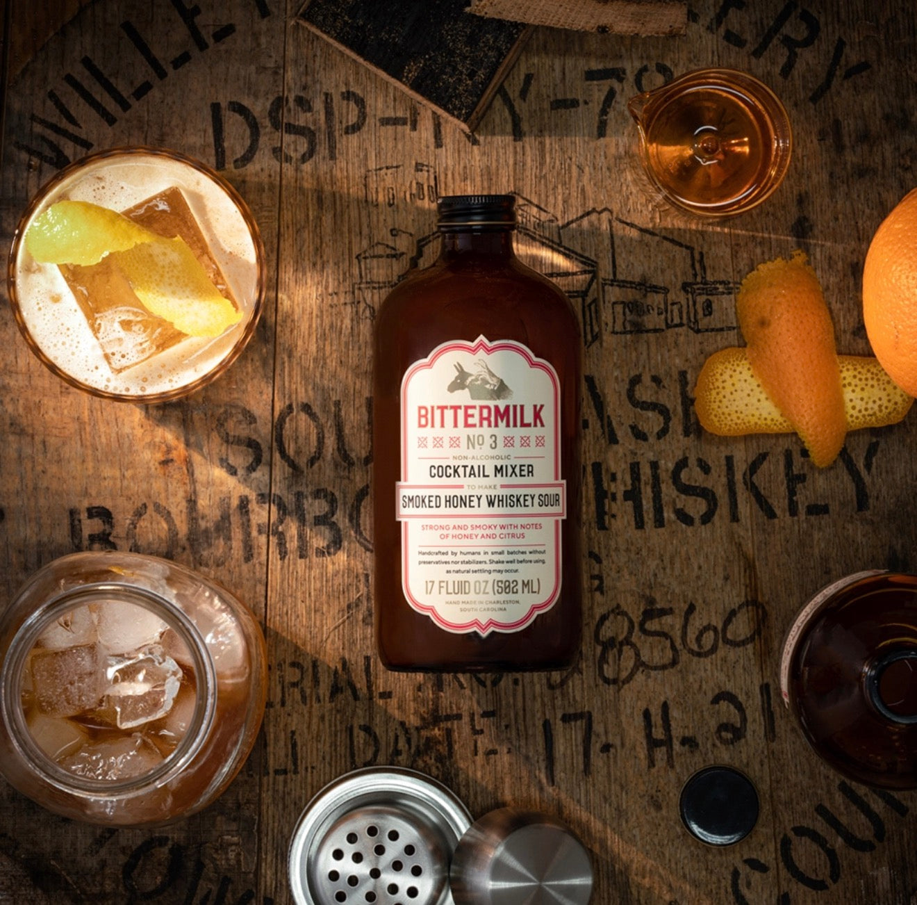 Bittermilk smoked honey whiskey sour mixer with cocktails citrus peel and bar tools on rustic bar surface