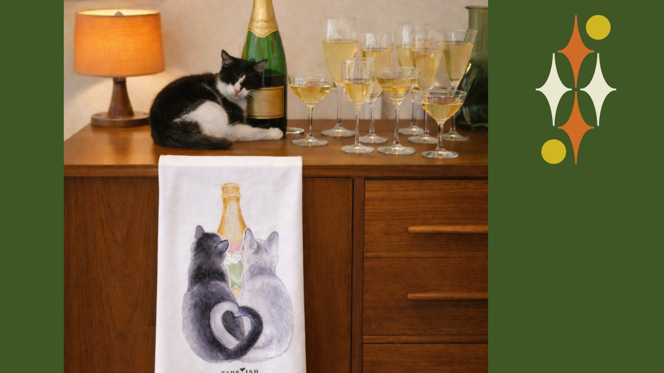 Champagne-themed cat cocktail tea towel hanging on a bar cabinet with champagne glasses and a black and white cat nearby