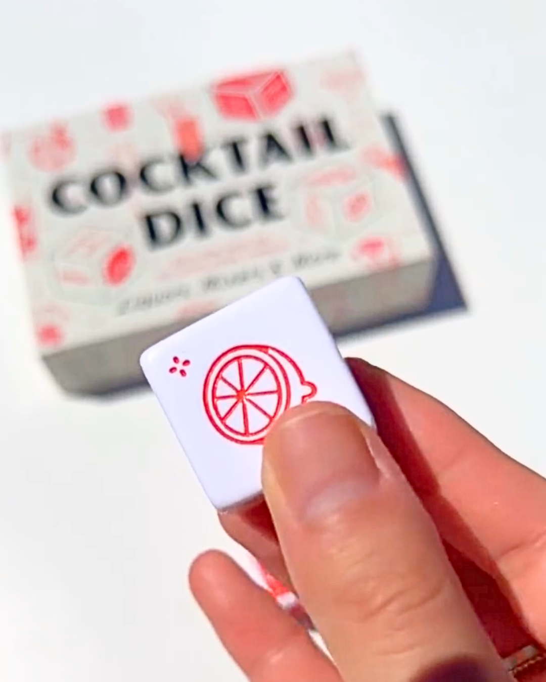 Close-up of a cocktail dice showing a citrus garnish icon, held in hand with the Cocktail Dice game box blurred in the background—fun gift for cocktail lovers.