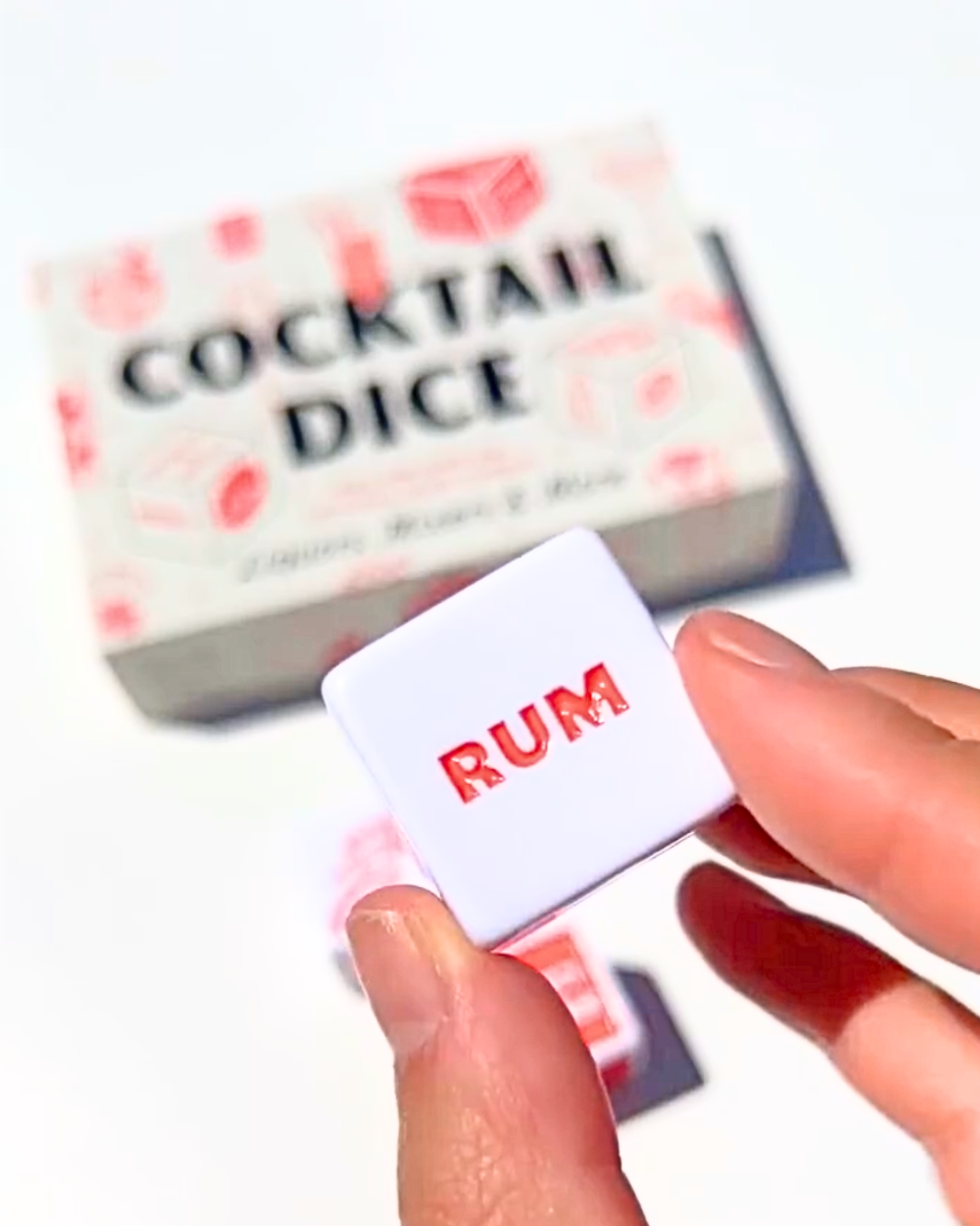 Close-up of a cocktail dice showing “Rum” in red lettering, held in hand with the Cocktail Dice game box softly blurred in the background—fun gift for cocktail lovers.