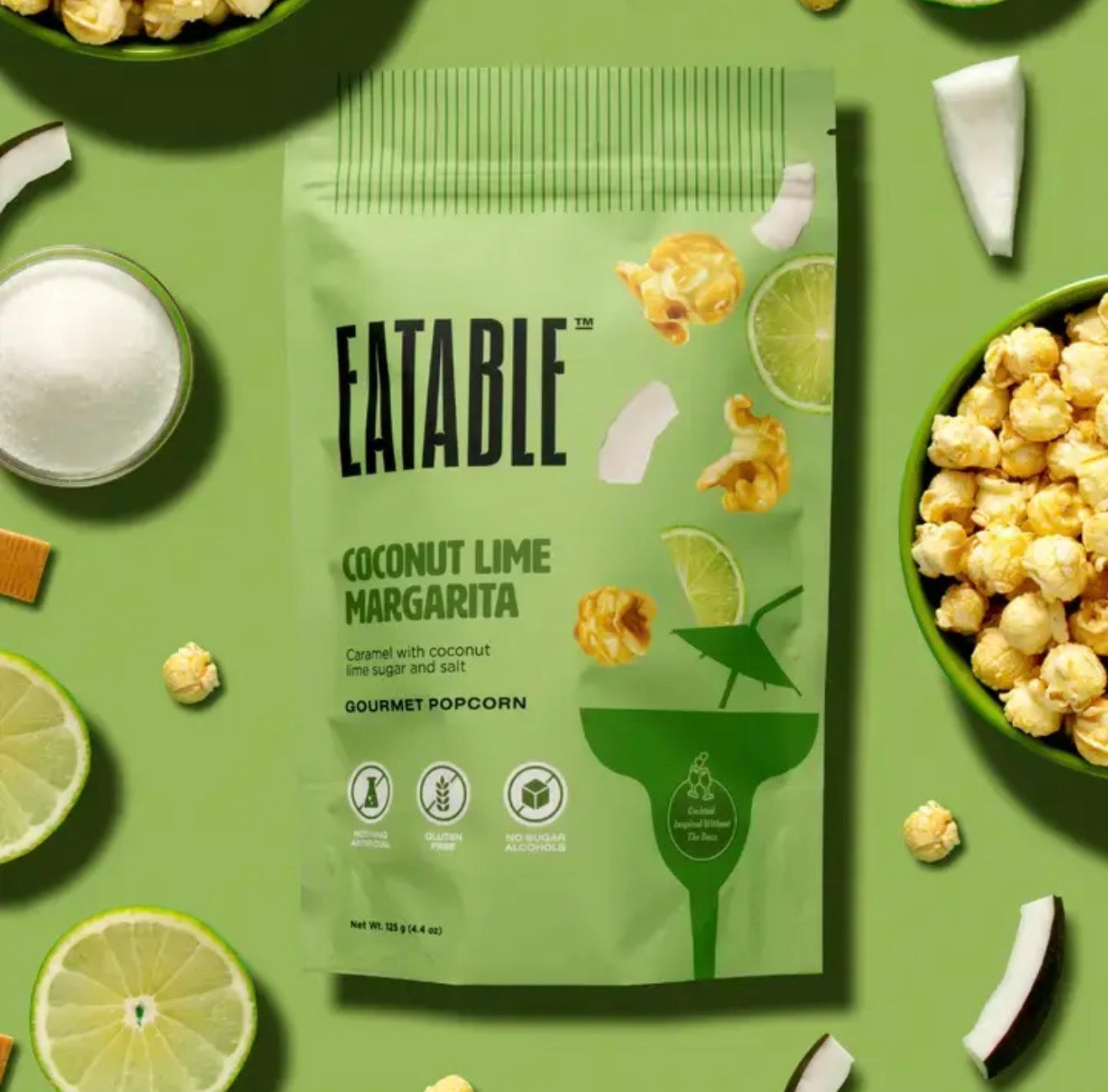 Flat lay of Coconut Lime Margarita popcorn bag with bowls of popcorn, lime slices, and coconut pieces