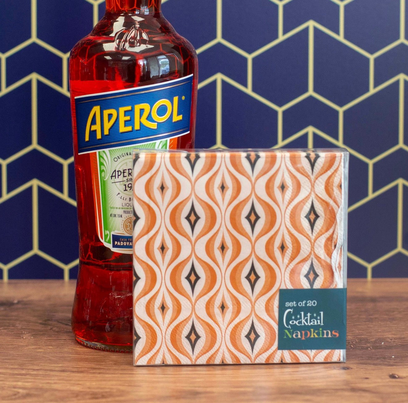 Orange retro cocktail napkins styled beside Aperol bottle on wood bar with geometric wallpaper