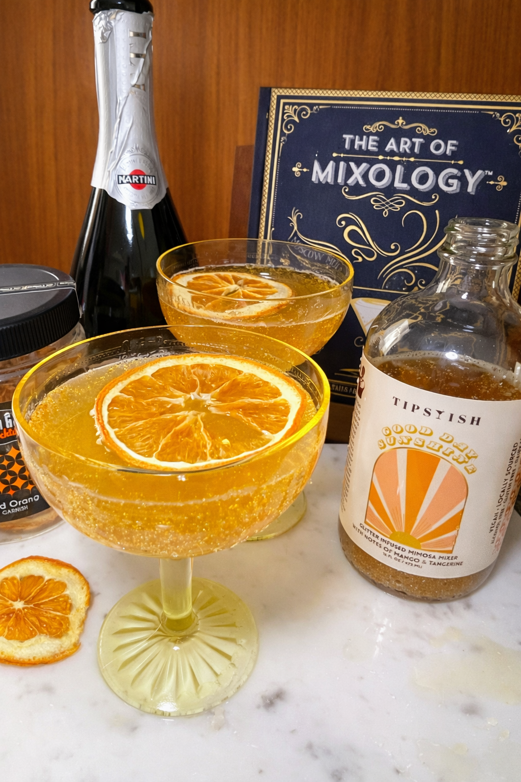 Sparkling mango tangerine mimosa with edible glitter served in coupe glasses with dried orange garnish and cocktail book