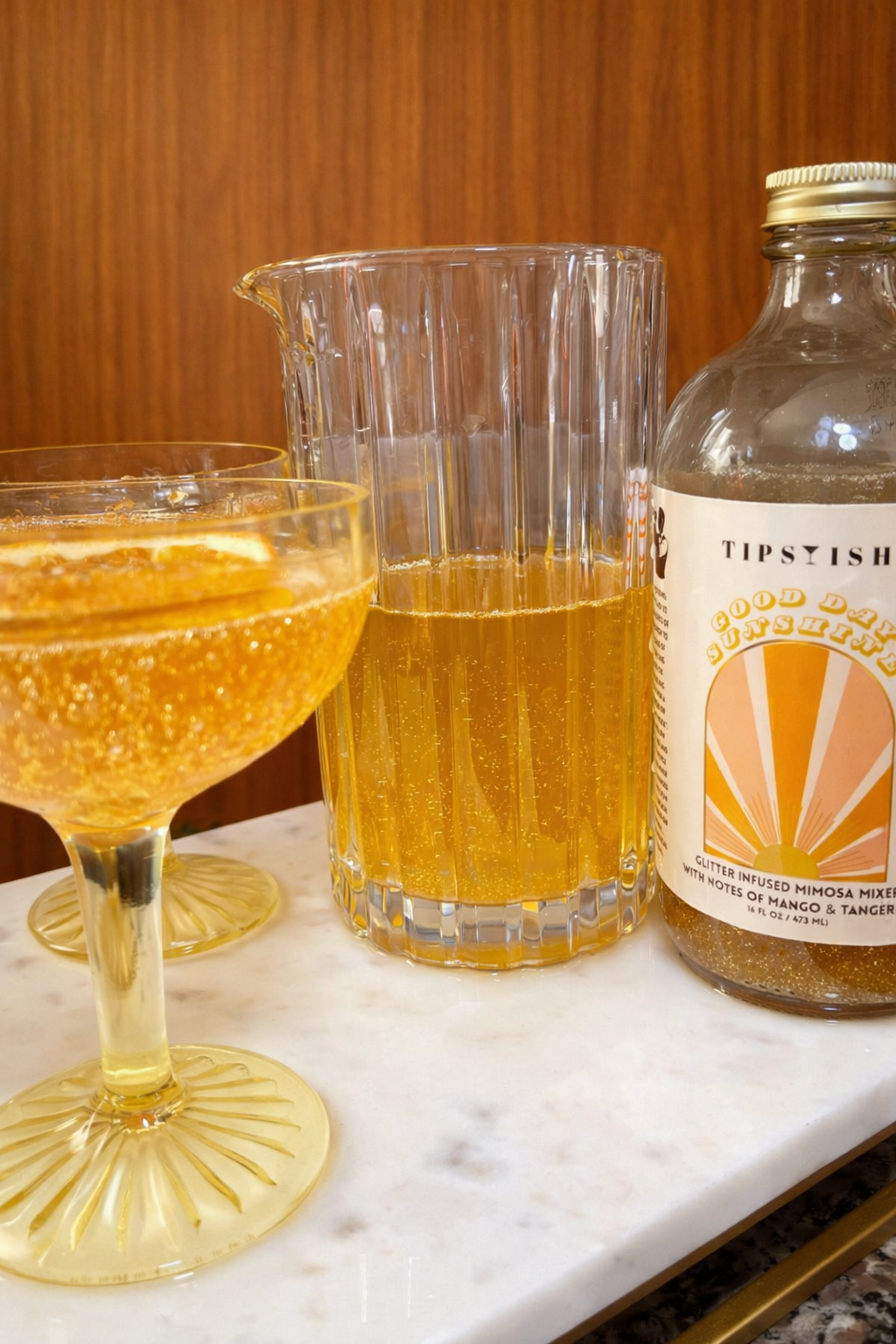 Pitcher and coupe glass filled with glitter mango tangerine mimosa next to Good Day Sunshine cocktail mixer bottle
