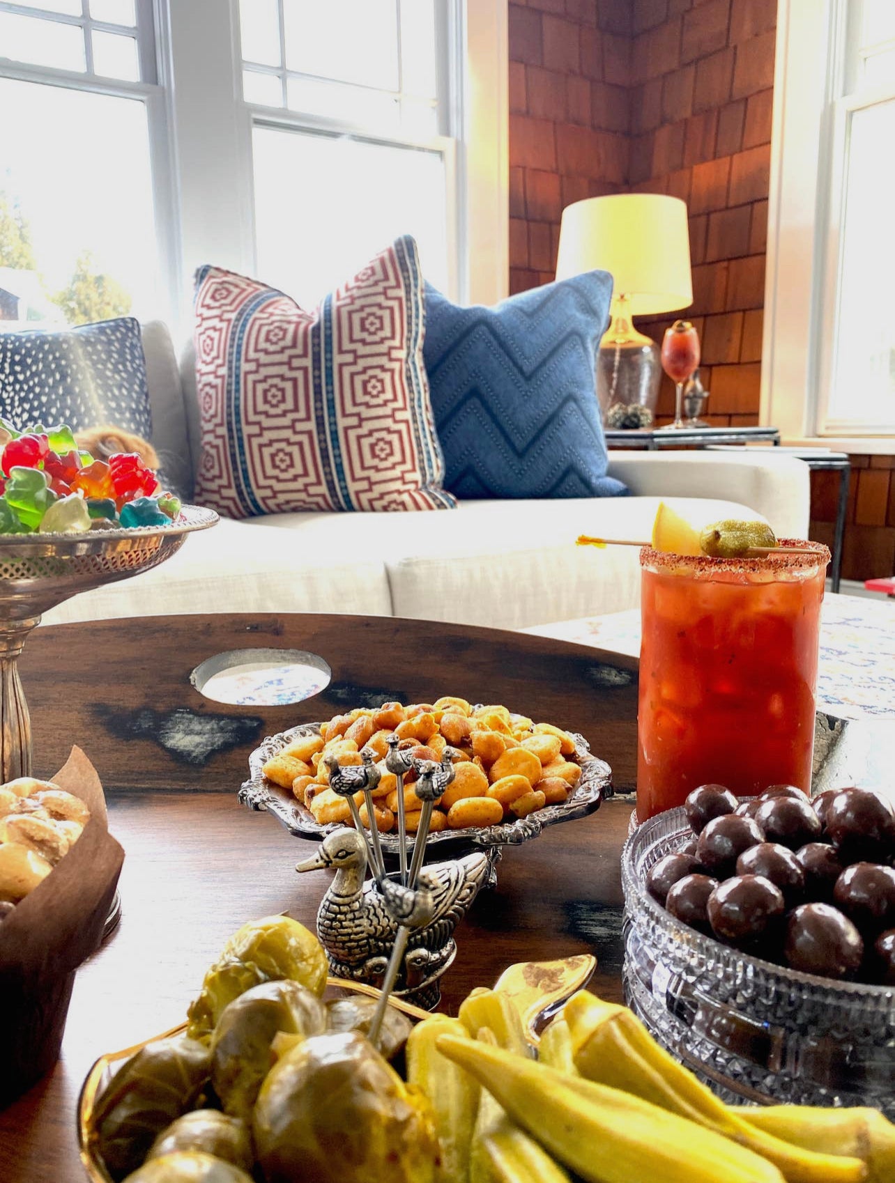 a bloody mary cocktail garnished with green olives sits on a coffee table in a family room surrounded by bowls of snacks