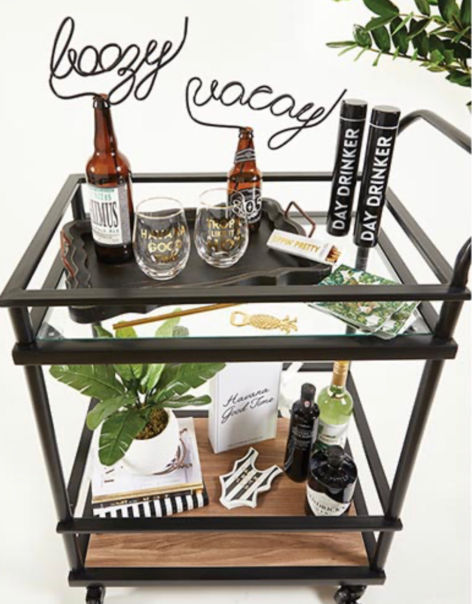 black plastic straw twisted into the word "boozy" at the top displayed sticking out of a beer bottle on a bar cart filled with glasses and bar tools
