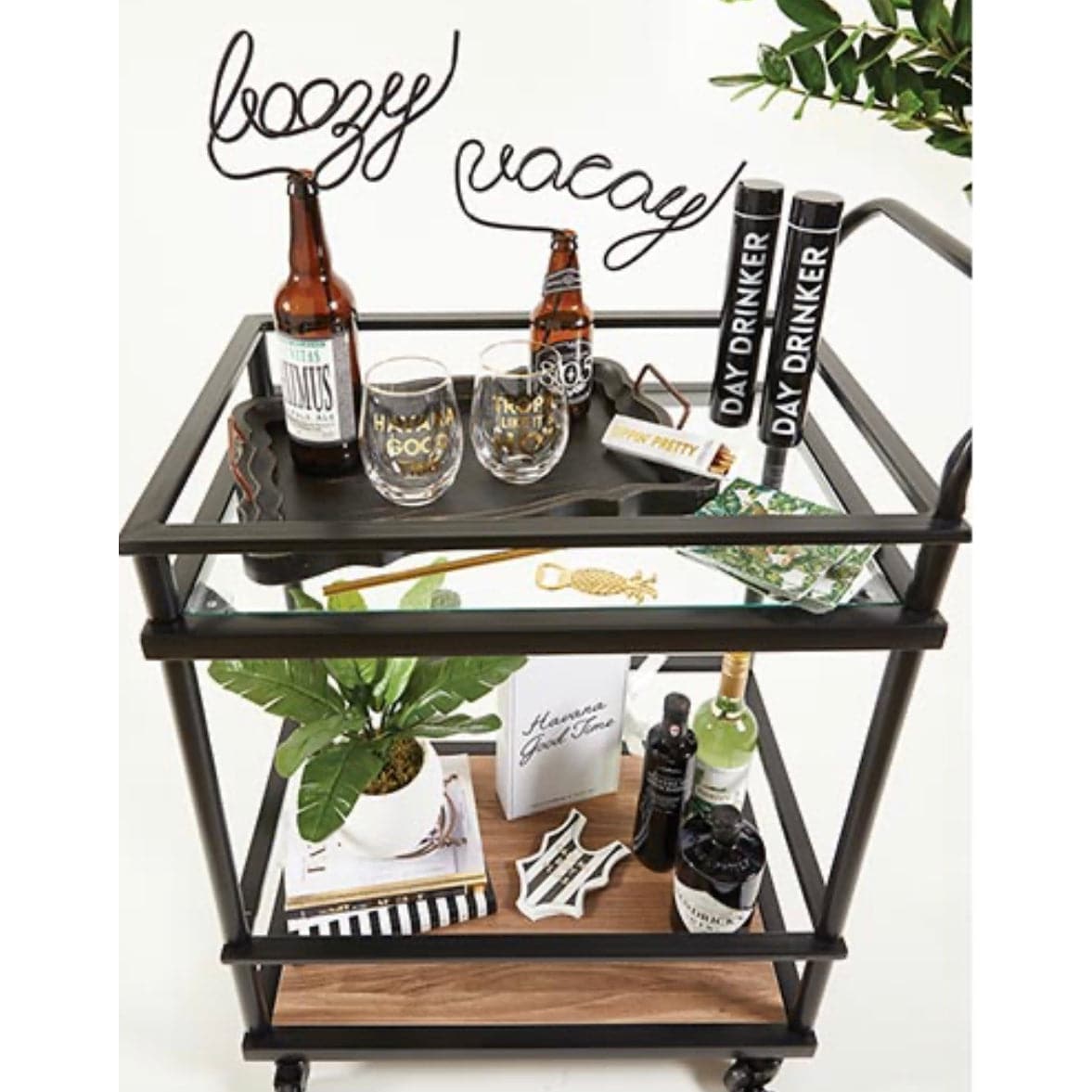 black plastic straw twisted into the word "boozy" at the top displayed sticking out of a beer bottle on a bar cart filled with glasses and bar tools