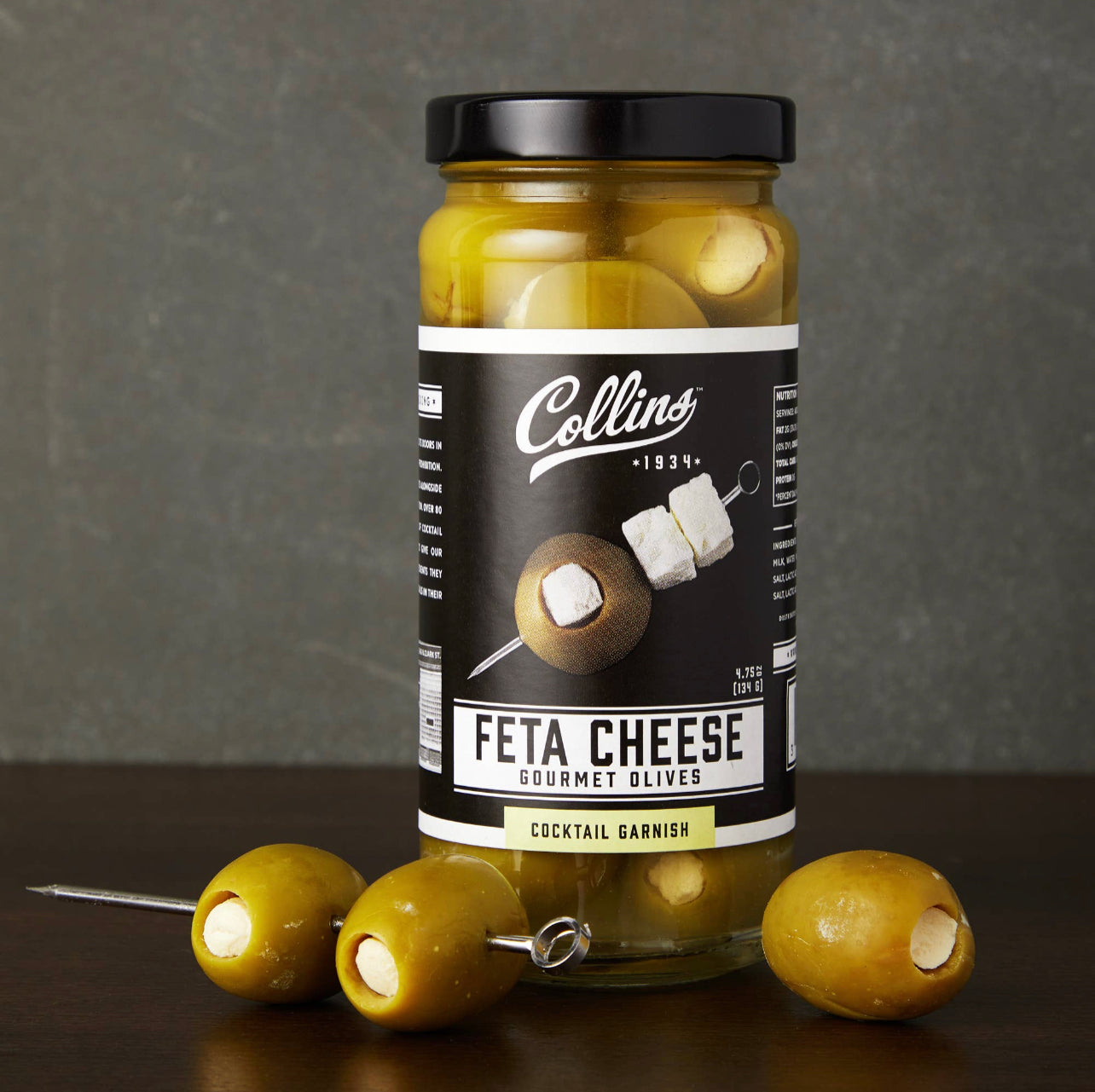 bottle of feta cheese gourmet olives cocktail garnish by Collins with examples of the actual olives displayed on a cocktail pick