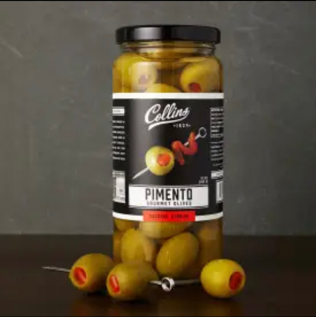 jar of pimento gourmet spanish olives by Collins displayed on a dark surface with some of the olives pictured on a cocktail pick