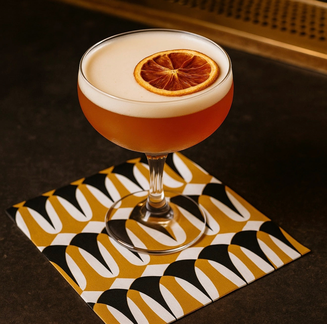 Coupe cocktail glass resting on black and amber geometric cocktail napkin on dark bar surface.