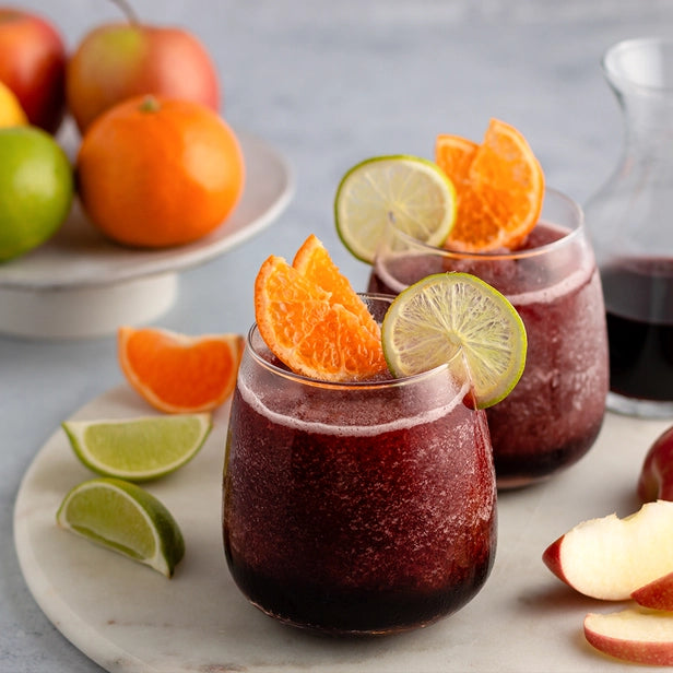 two stemless wine glasses containing frozen sangria slushies that are garnished with fresh limes, oranges, and apples