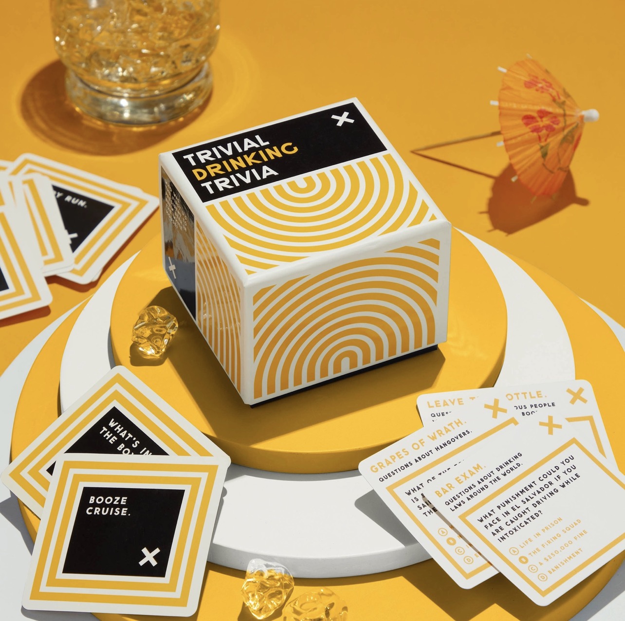 Trivial Drinking Trivia game box styled with trivia cards and cocktail accents on a bright yellow surface, a fun party game gift for cocktail lovers.