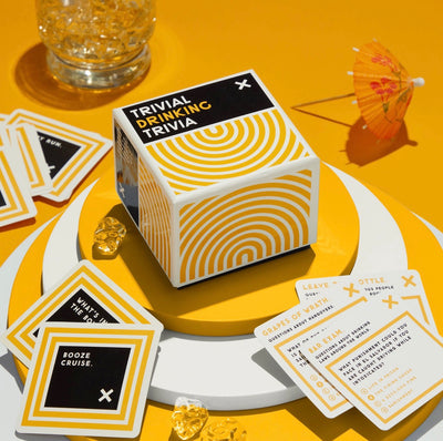 Trivial Drinking Trivia game box styled with trivia cards and cocktail accents on a bright yellow surface, a fun party game gift for cocktail lovers.