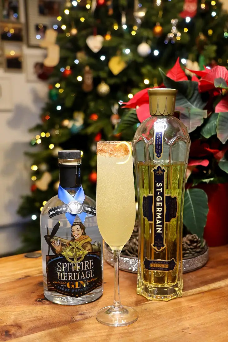 cocktail in a champagne flute featuring the bronze sugar dust rimming the glass accompanied by liquor bottles with a holiday scene in the background