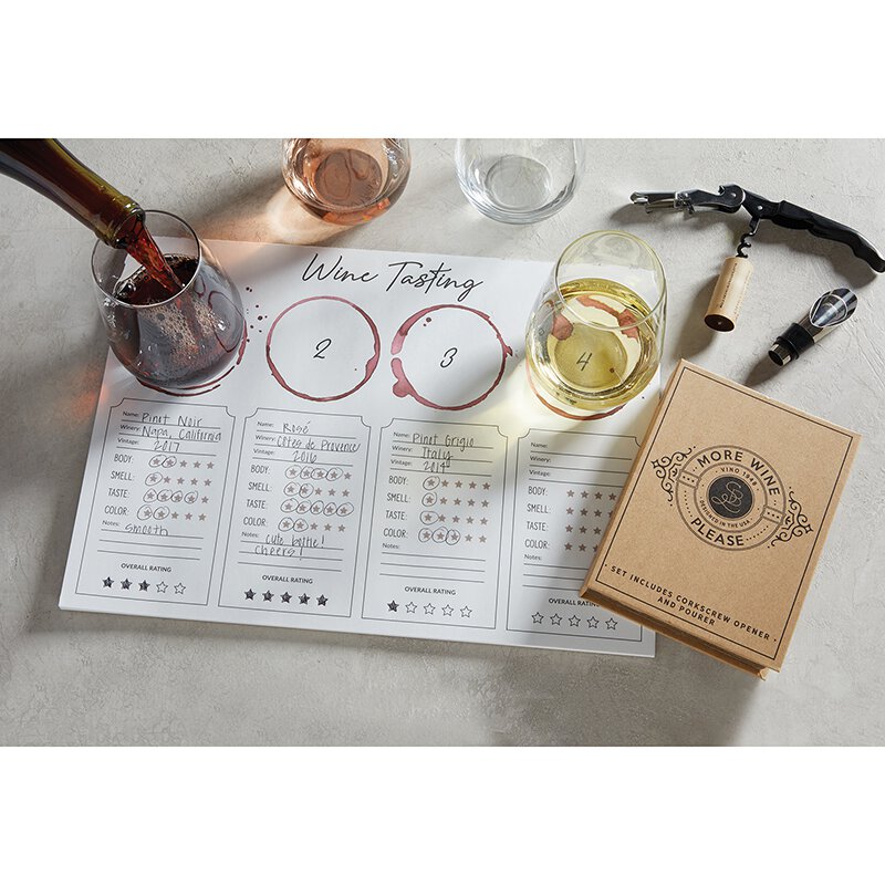"More Wine Please" craft gift box that includes a corkscrew opener and pourer displayed with a wine tasting placemat and wine glasses filled with different types of wine