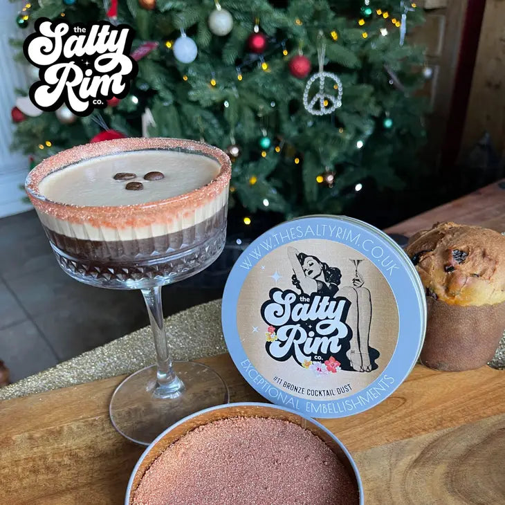 Container of bronze cocktail dust to rim glasses made by The Salty Rim displayed with an espresso martini with the bronze dust rimming the glass and a holiday scene in the background
