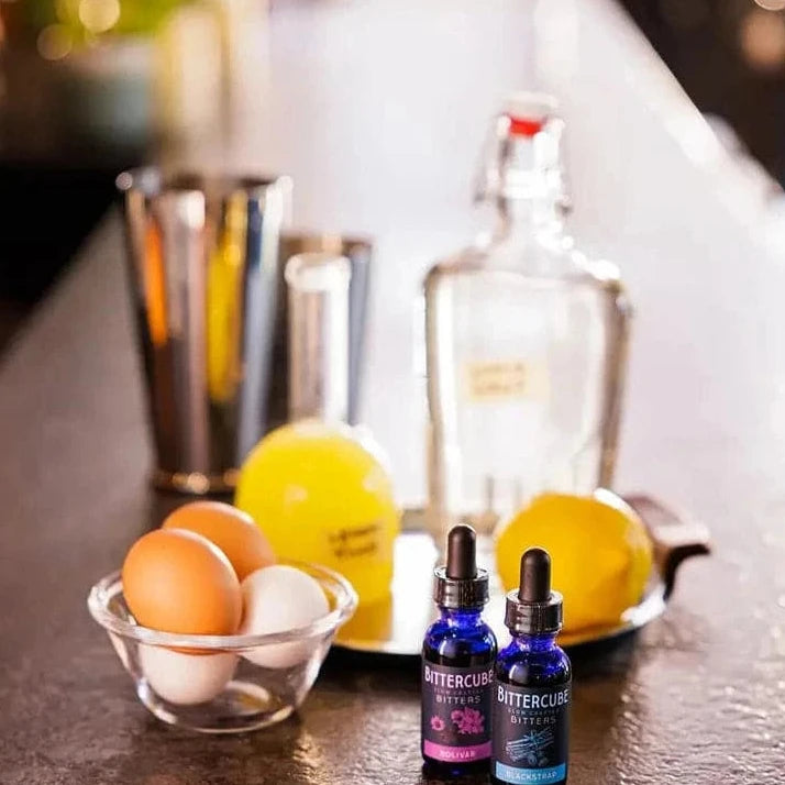 Bottles of cocktail bitters displayed with spirits, glasses, citrus and eggs 