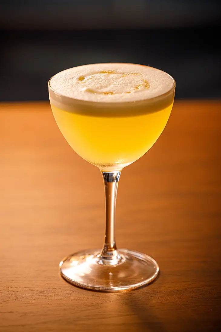 a golden cocktail in a stemmed glass with a white soft top displayed on a wooden surface