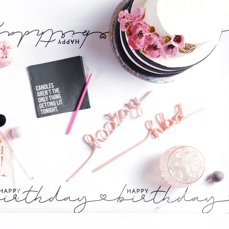 5" x 5" black square paper napkin with the words "Candles Aren't The Only Thing Getting Lit Tonight" in white, while the words "getting lit" stand out in the color pink. The napkins are displayed on a table decorated for a party with a cake, birthday table runner, and pink birthday straws