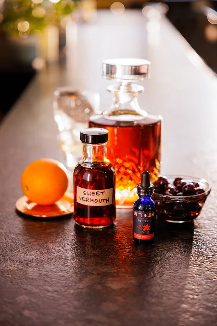 1 oz bottle with dropper of cherry bark vanilla bitters by Bittercube displayed with a whiskey decanter and vermouth, orange, and cherries on a bar top