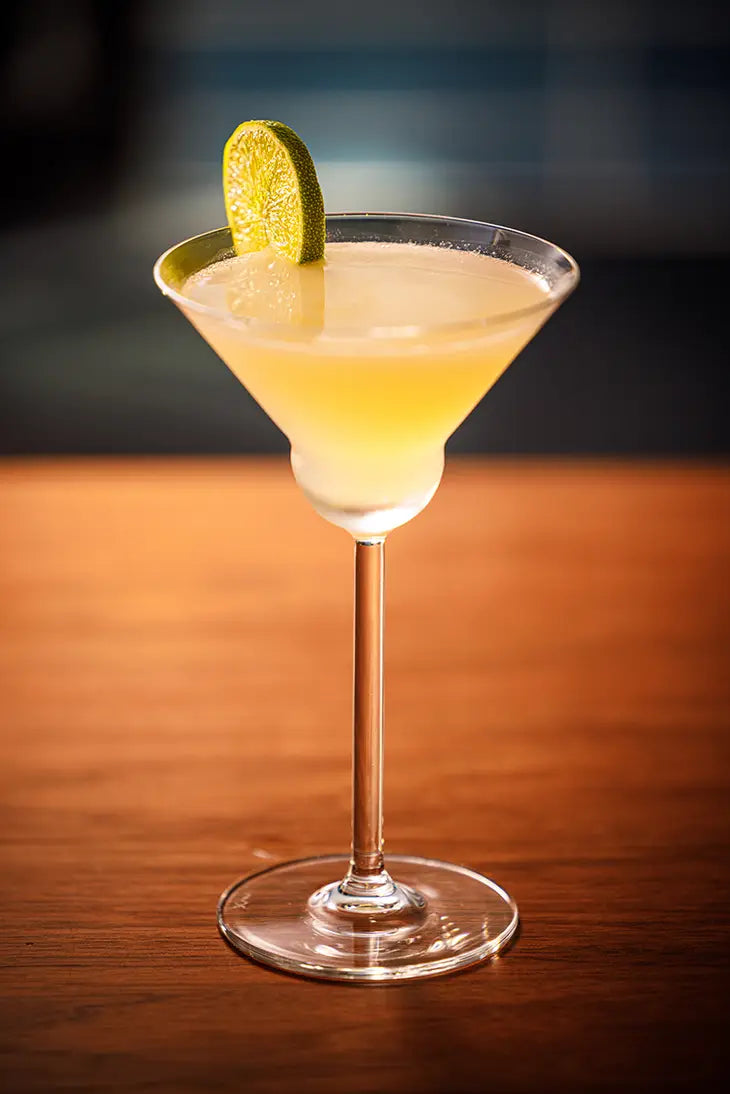 golden cocktail in a clear martini glass garnished with lime sitting on a wooden surface