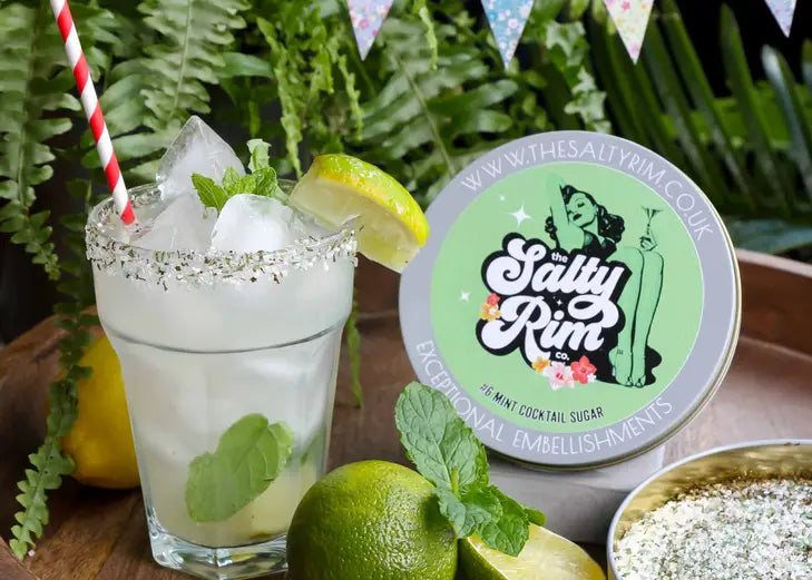 tin of mint cocktail sugar by The Salty Rim Co. displayed alongside a mojito rimmed with the sugar and garnished with lime; the bottom half of the tin is open for display of the mint green color of the sugar