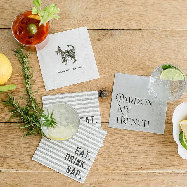 5" x 5" white square cocktail napkin featuring a modern sketch of a pup in black with the phrase "hair of the dog" printed underneath displayed with different styles of napkins and cocktails on a wooden surface
