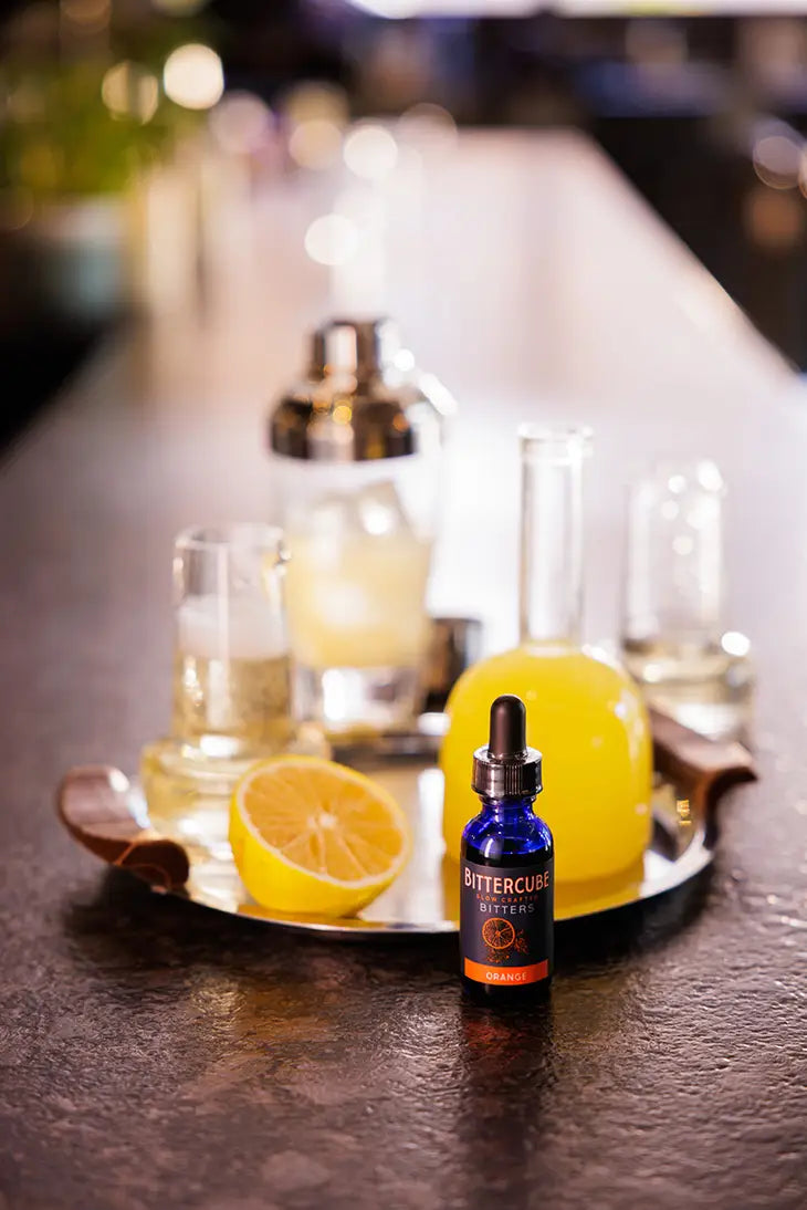 1 oz bottle with dropper of Orange bitters by Bittercube displayed in fron of a silver bar tray with glasses, cocktail shaker, and fresh orange on a bar top