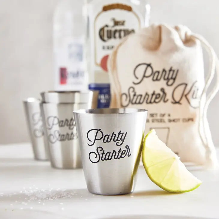 set of four stainless steel shot cups engraved with the words "party starter" displayed with the linen storage bag, liquor, and fresh lime